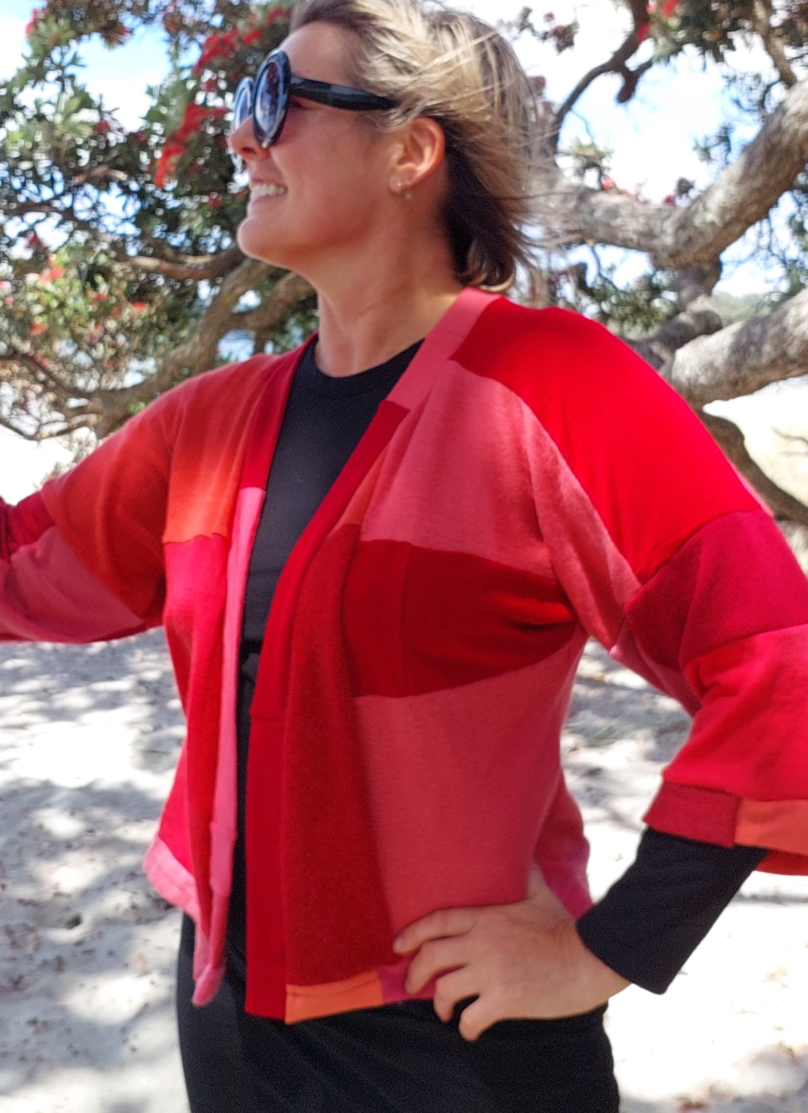 Side profile view of a client on a coastal beach wearing a custom upcycled merino wool short kimono in bright red and coral patchwork panels, showcasing the relaxed sleeve drape.