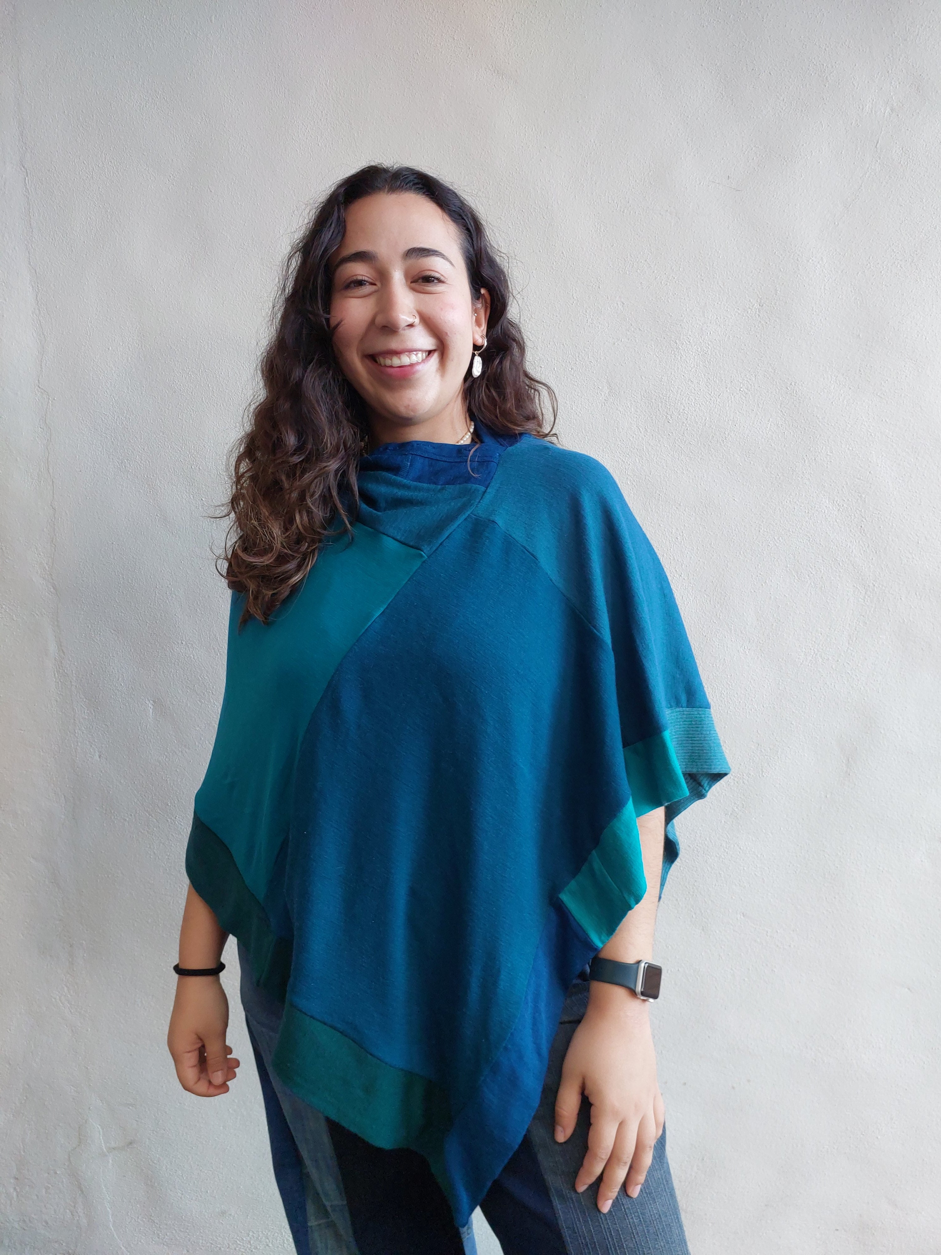Front view of a model outdoors wearing a handcrafted upcycled merino wool poncho in a vibrant aqua and teal patchwork design, showing the asymmetric drape.