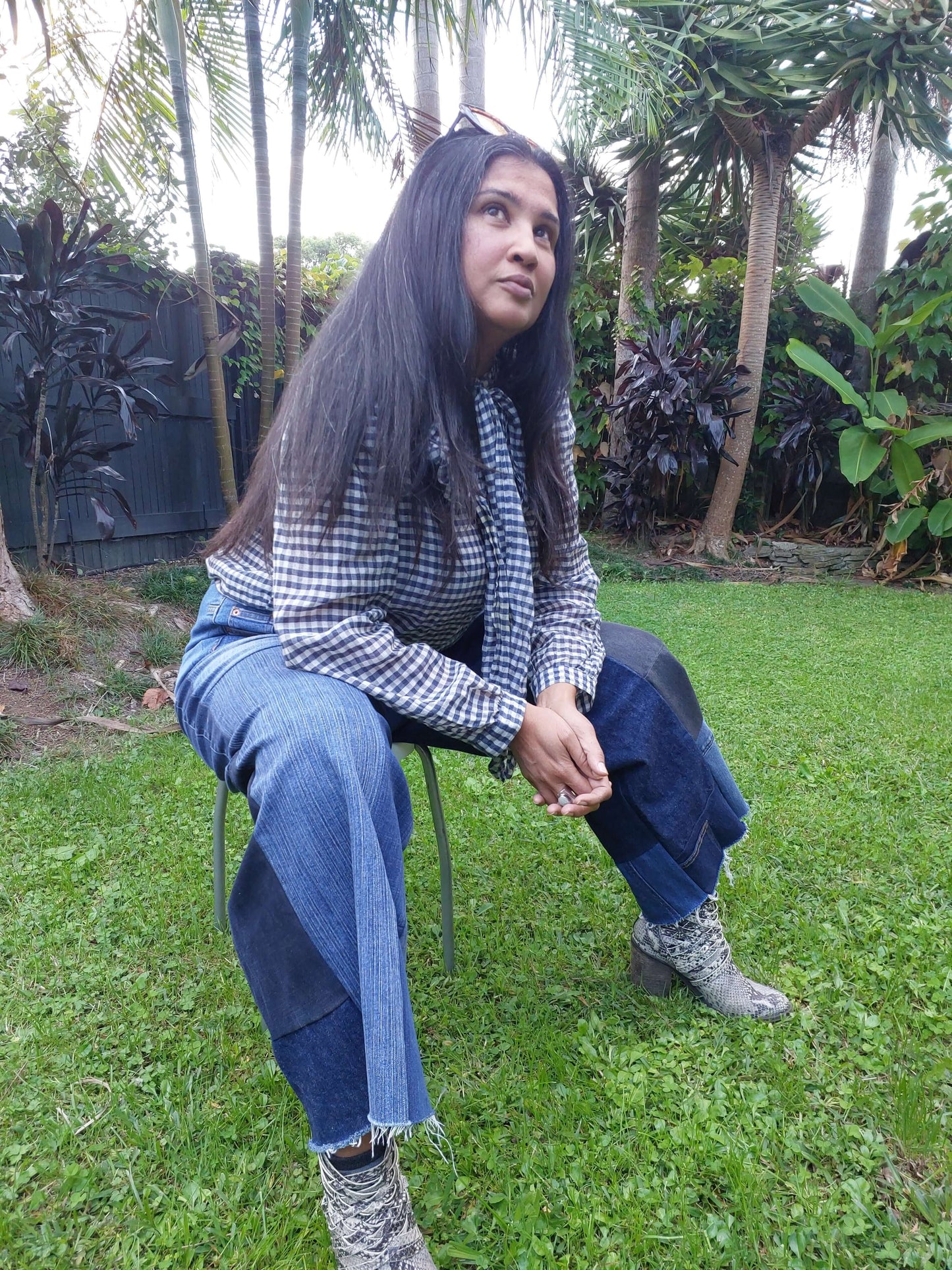 Lifestyle shot of a model wearing custom-size upcycled patchwork jeans by Heke Design, seated in a lush Waiheke garden setting.