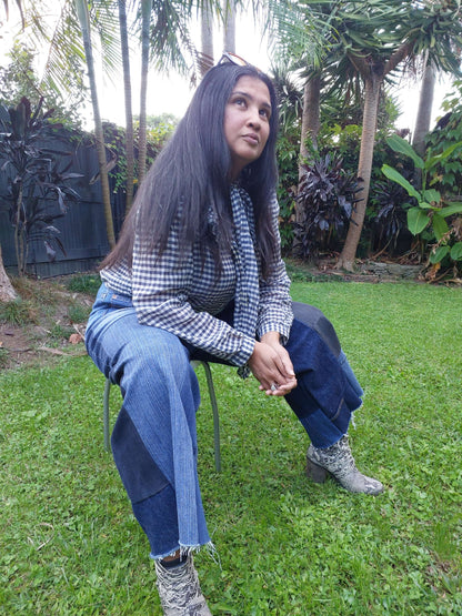 Lifestyle shot of a model wearing custom-size upcycled patchwork jeans by Heke Design, seated in a lush Waiheke garden setting.