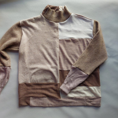 A color-blocked knit sweater laid flat on a light gray fabric background. The sweater features panels of taupe, light brown, cream, and white, with a ribbed mock neck and cuffs.