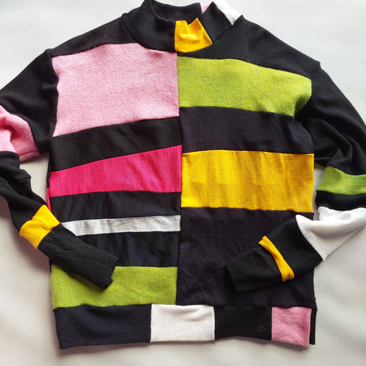 A black knit sweater with large color-blocked panels in pink, lime green, yellow, white, and a vertical magenta stripe, laid flat on a white fabric background.